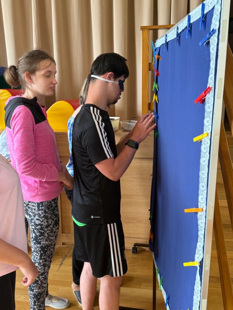 Grupa osób stoi przed tablicą. Chłopiec z zakrytymi opaską oczami dotyka kolorowych spinaczy przyczepionych do obrzeży tablicy