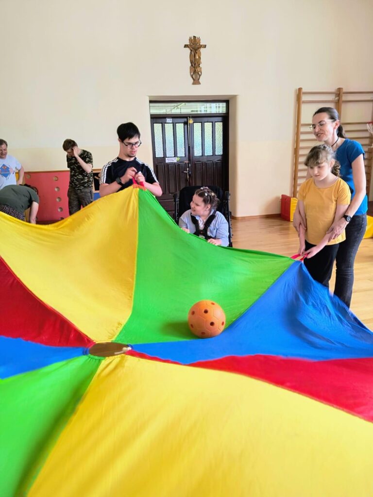 Grupa osób trzyma za uchwyty dużą kolorową chustę do zabaw zespołowych na której znajduje się piłka.
