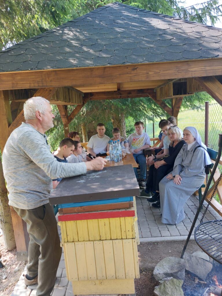 Grupa osób siedzi pod drewniana altaną, słuchając starszego mężczyzny, który prowadzi prezentację. Na stole stoją butelki z wodą, a w okół panuje spokojna, leśna atmosfera.