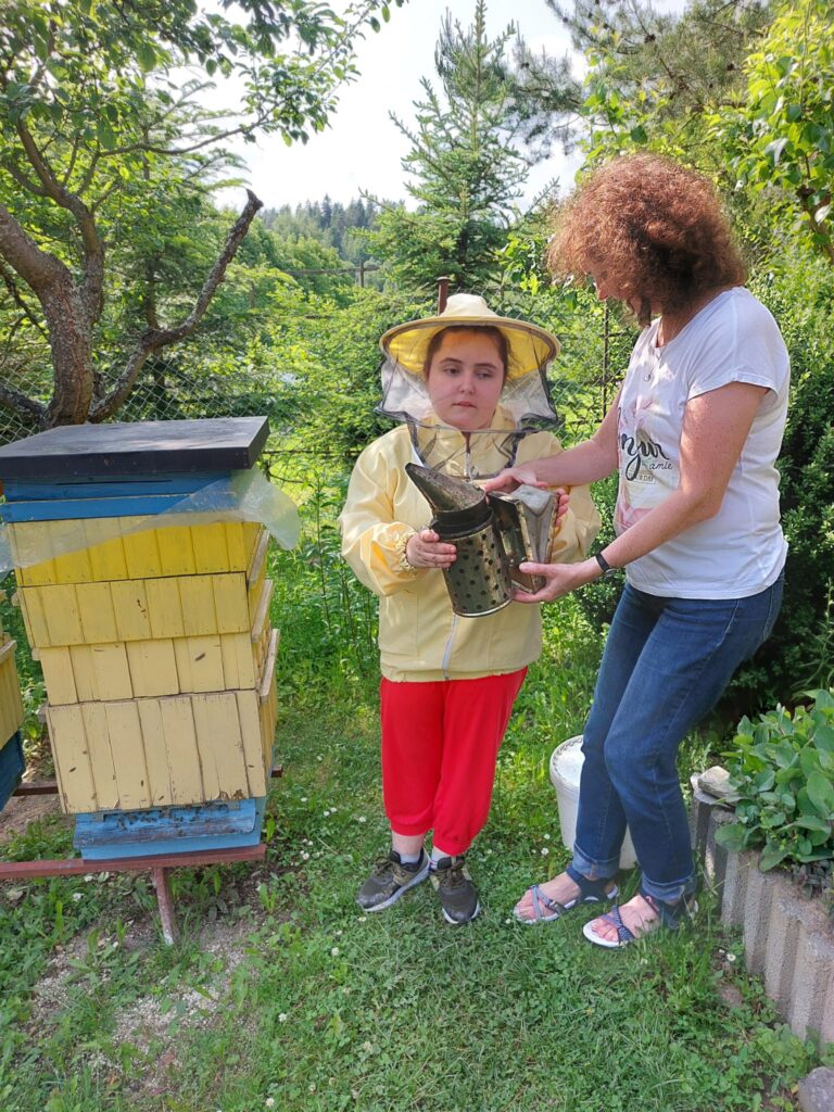 Dziewczynka ubrana w strój pszczelarza stoi obok ula obok niej znajduje się kobieta. Obie trzymają w rękach podkurzacz.