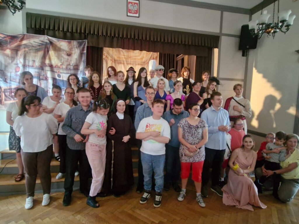 Zdjęcie grupowe przedstawiające uczestników przedstawienia teatralnego. W centrum osoby z różnymi niepełnosprawnościami, wokół nich aktorzy w kostiumach historycznych. Scena udekorowana malowanymi tłami.