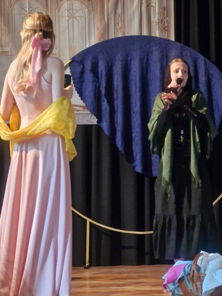 Scena teatralna: kobieta w różowej sukni z żółtym szalem stoi tyłem, naprzeciw niej kobieta w czarnym stroju z zieloną chustą śpiewa lub mówi do mikrofonu. W tle pałacowa dekoracja.