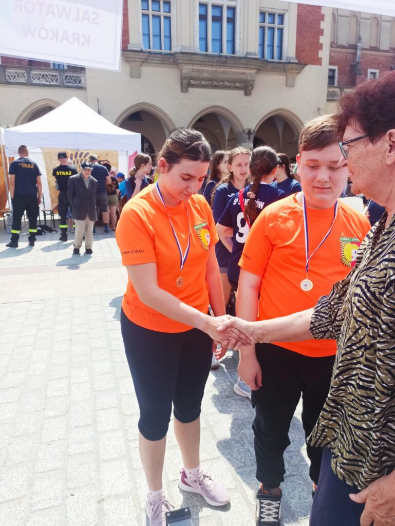 Młoda kobieta i mężczyzna w pomarańczowych koszulkach z medalami na szyi przyjmują gratulacje od starszej kobiety. 