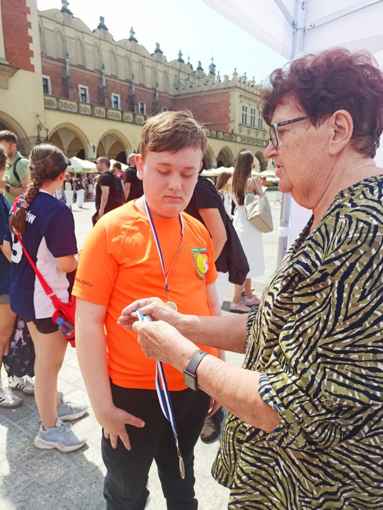 Chłopiec w pomarańczowej koszulce po dekoracji medalem, w tle krakowski rynek.