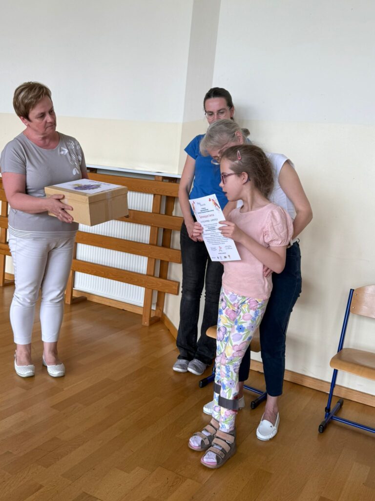 Dziewczynka otrzymuje dyplom i prezent, stojąc z dwiema dorosłymi kobietami w sali szkolnej; jedna z kobiet wręcza jej pudełko z nagrodą.