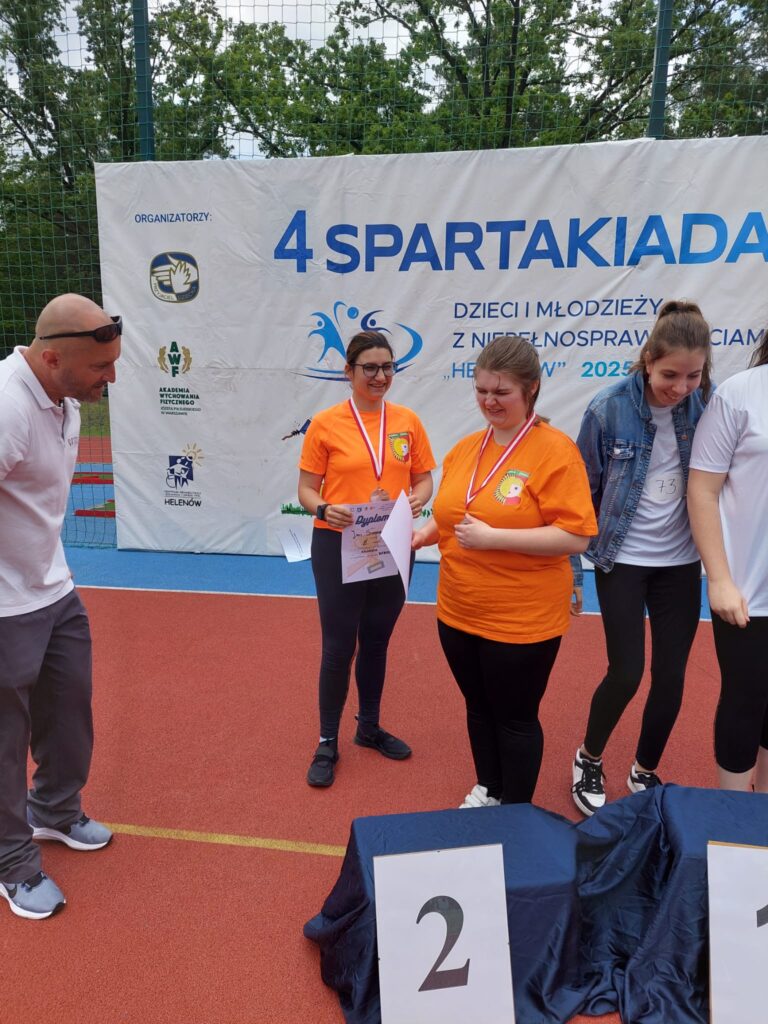 Dwie dziewczyny w pomarańczowych koszulkach stoją koło podium z medalami i dyplomami podczas zawodów sportowych.