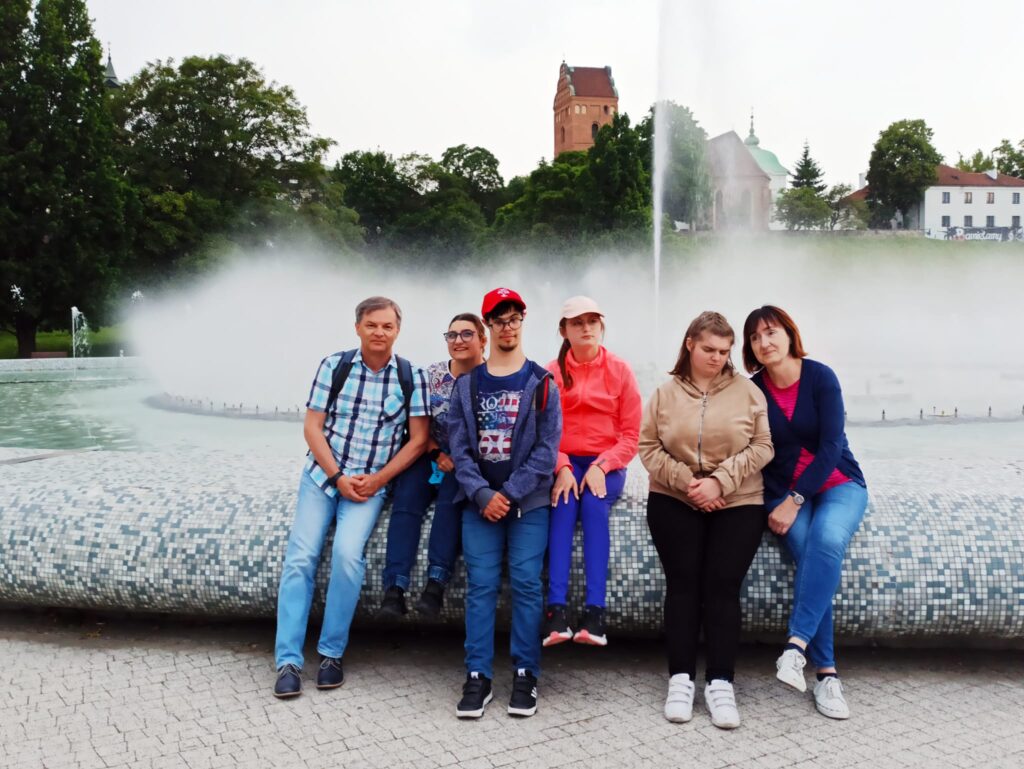 Młodzież z opiekunami siedzą na obrzeżu fontanny.