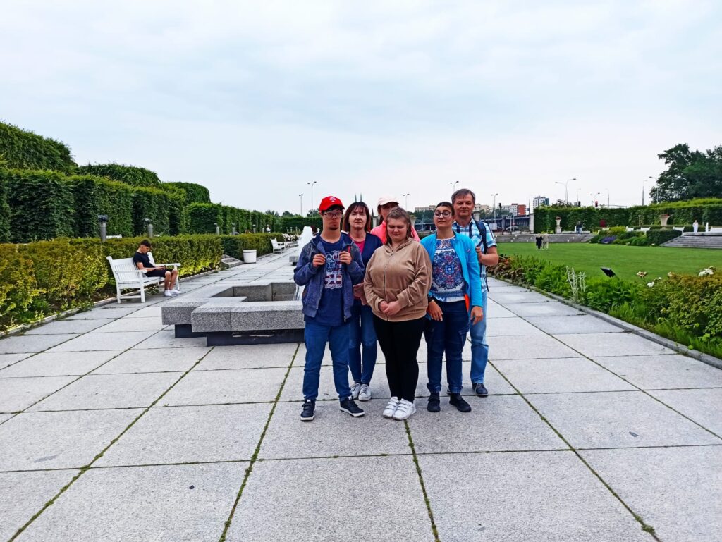 Grupa osób na deptaku spacerowym, w tle fontanna, ławki i dużo zieleni.