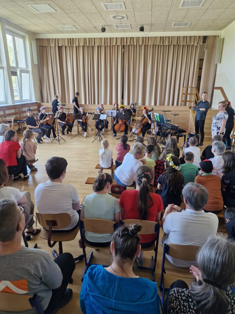 Widok sali z publicznością siedzącą na krzesłach, przed sceną młodzi muzycy z instrumentami smyczkowymi i keyboardem przygotowani do występu.