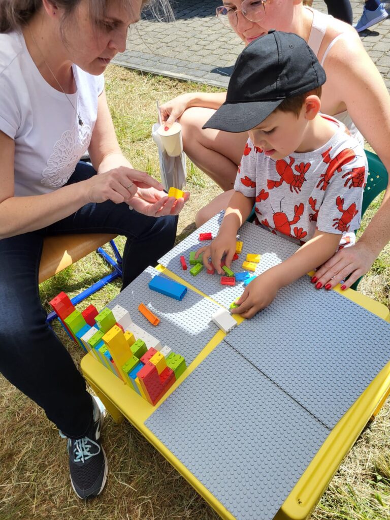 Dziecko bawi się klockami "Lego Braille" przy stole na zewnątrz, w towarzystwie dwóch dorosłych.