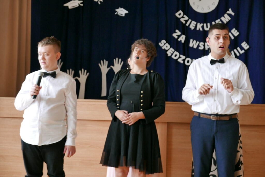 Trójka młodych ludzi ubrana elegancko, występująca na scenie z mikrofonami, na tle dekoracji z napisem „Dziękujemy za wspólnie spędzone chwile”.