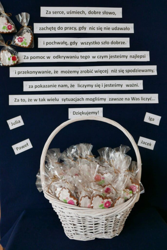 Biały koszyk wypełniony kolorowo dekorowanymi pierniczkami zapakowanymi w celofan, ustawiony na stole przed granatowym materiałem, a za nim napis dziękczynny.