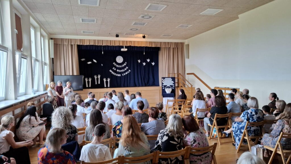 Sala gimnastyczna z widownią skierowaną na scenę, na której odbywa się uroczystość zakończenia roku szkolnego.