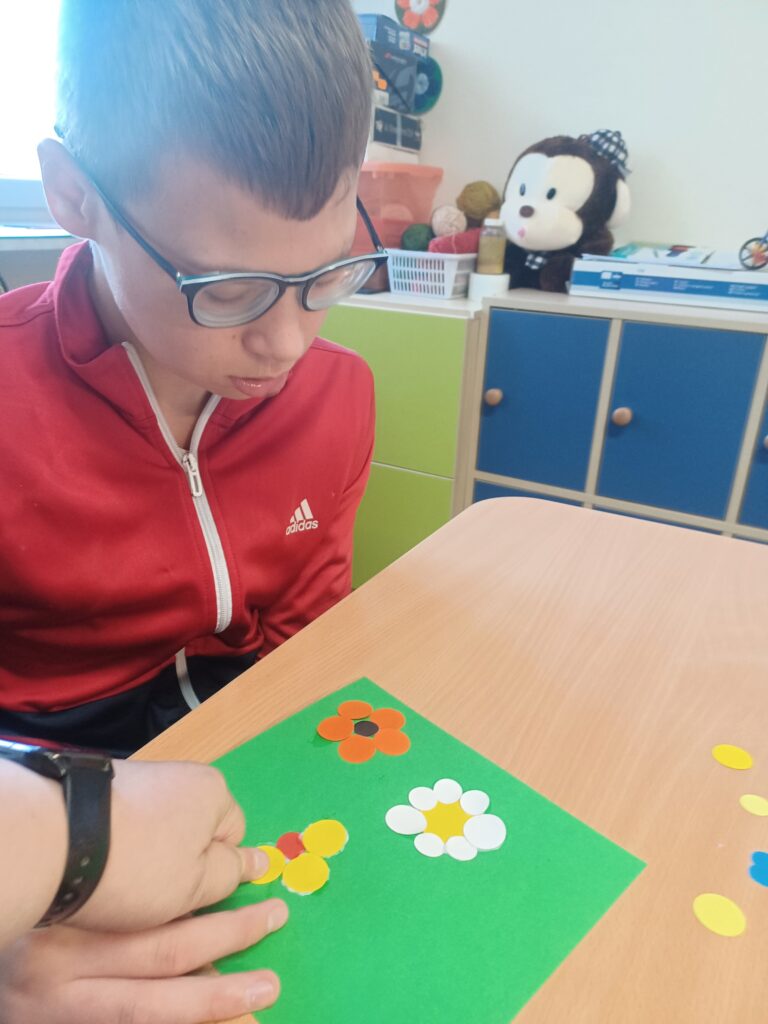 Chłopiec w czerwonej bluzie i okularach nakleja na kartkę żółte kółeczko. 