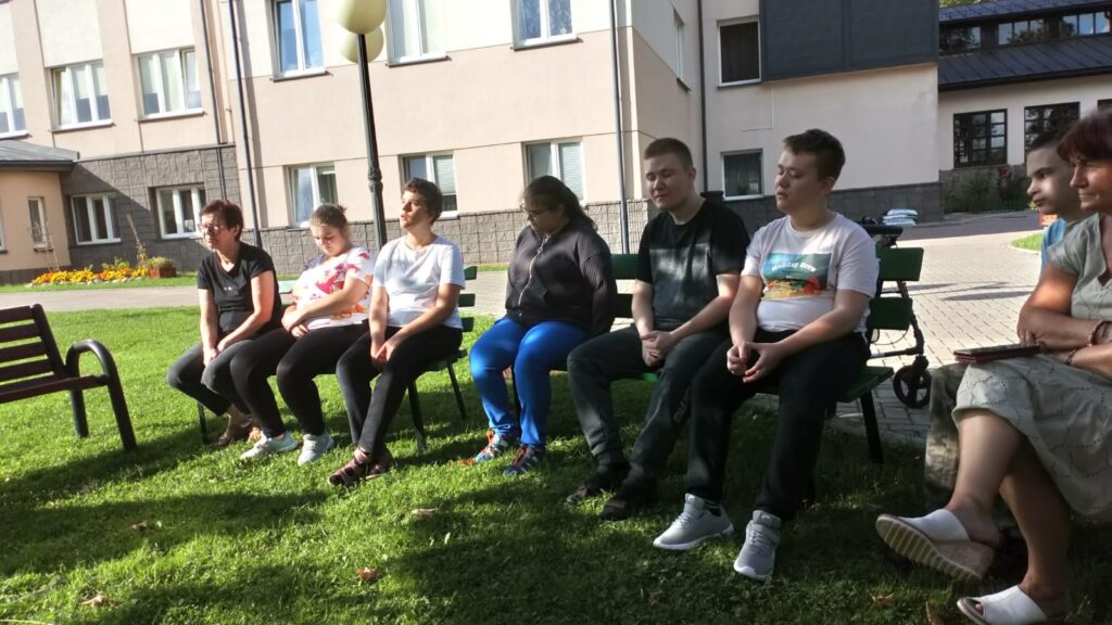 Grupa młodzieży i dorosłych siedzą na ławkach przed szkołą i uważnie czegoś słuchają.