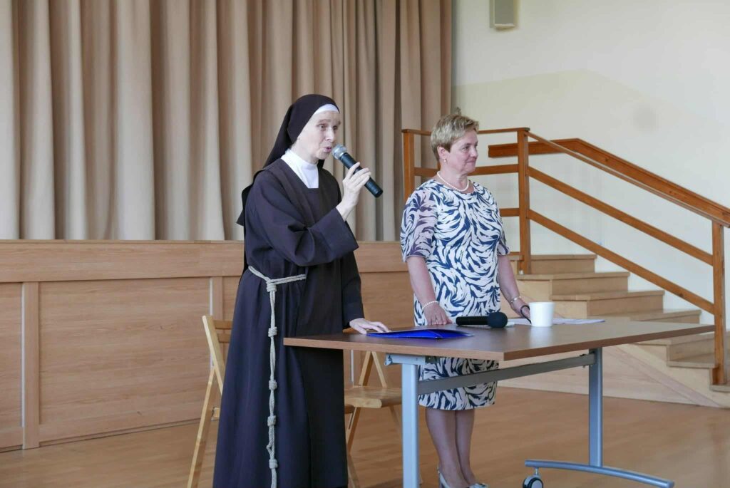 Siostra zakonna w brązowym habicie oraz kobieta  stoją przy stole. Siostra przemawia przez mikrofon.