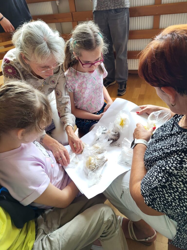 Dwie kobiety z dziewczynkami sprawdzają zawartość woreczków nalepionych na dużą kartkę.