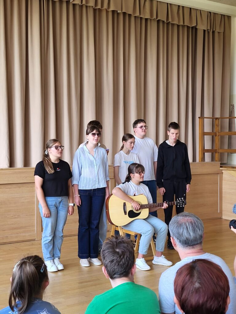 Grupa młodych osób stoi przy scenie, a jedna dziewczyna siedzi z gitarą i gra. Z przodu widać osoby fotografujące i nagrywające występ.