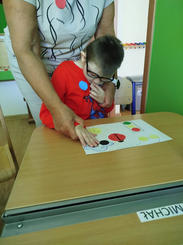 Chłopiec w czerwonej bluzie i okularach nakleja na kartce z pomocą nauczyciela czarne kółko.