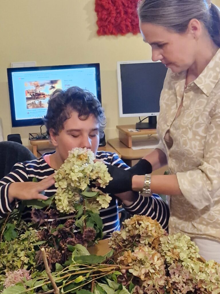 Kobieta pomaga osobie siedzącej przy stole wąchać bukiet hortensji. Na stole leżą różne rośliny. W tle widoczne są komputery i monitor z filmem przyrodniczym.