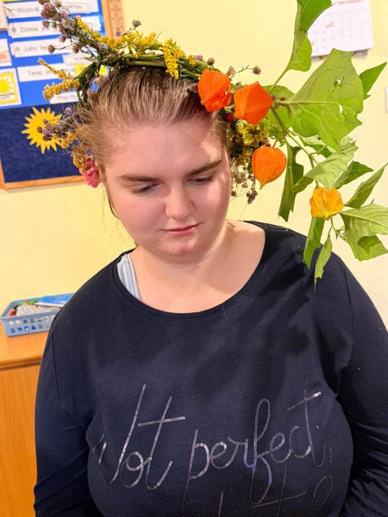 Dziewczyna w ciemnej bluzce, ma na głowie wianek z żółtych i pomarańczowych kwiatów oraz lisci.