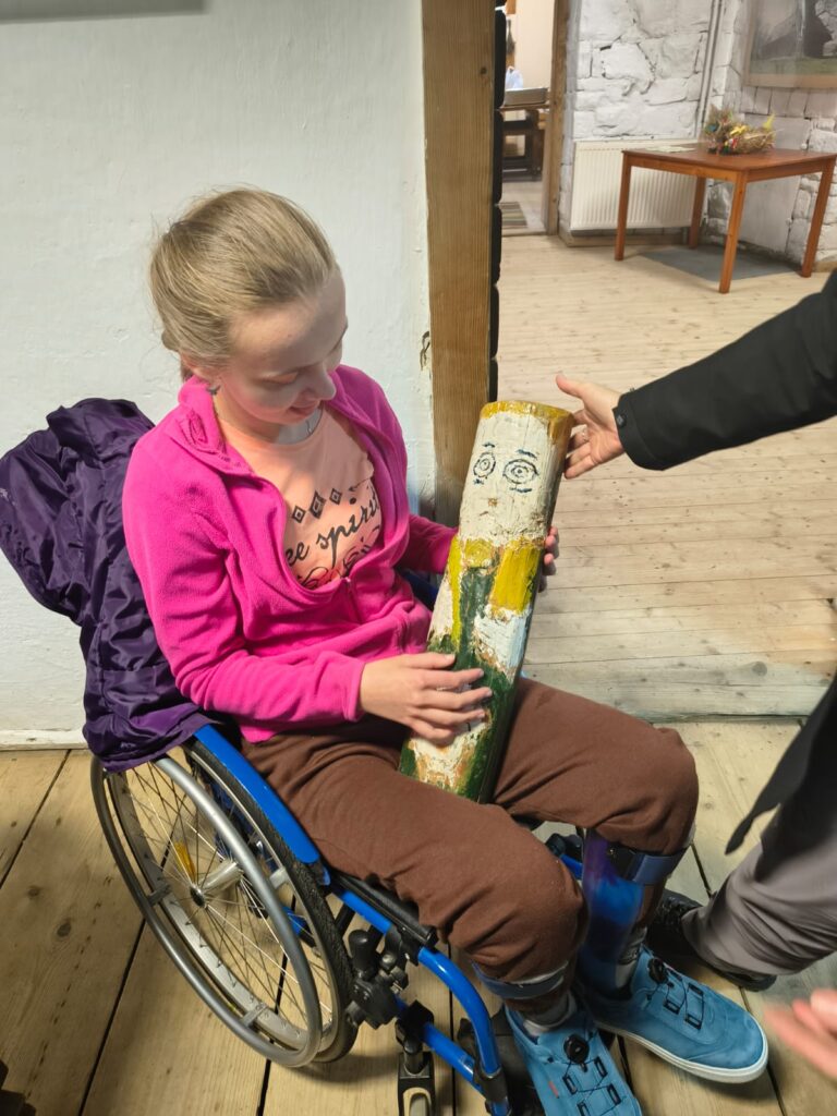 Dziewczyna na wózku inwalidzkim trzyma w rękach drewnianą pomalowana rzeźbę.