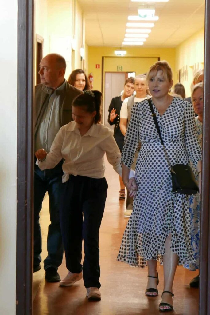 Grupa dorosłych i dzieci przechodzi korytarzem szkolnym. Na pierwszym planie kobieta w sukience w grochy trzyma za rękę dziewczynkę ubraną na biało i granatowo. Inni dorośli i dzieci idą za nimi.