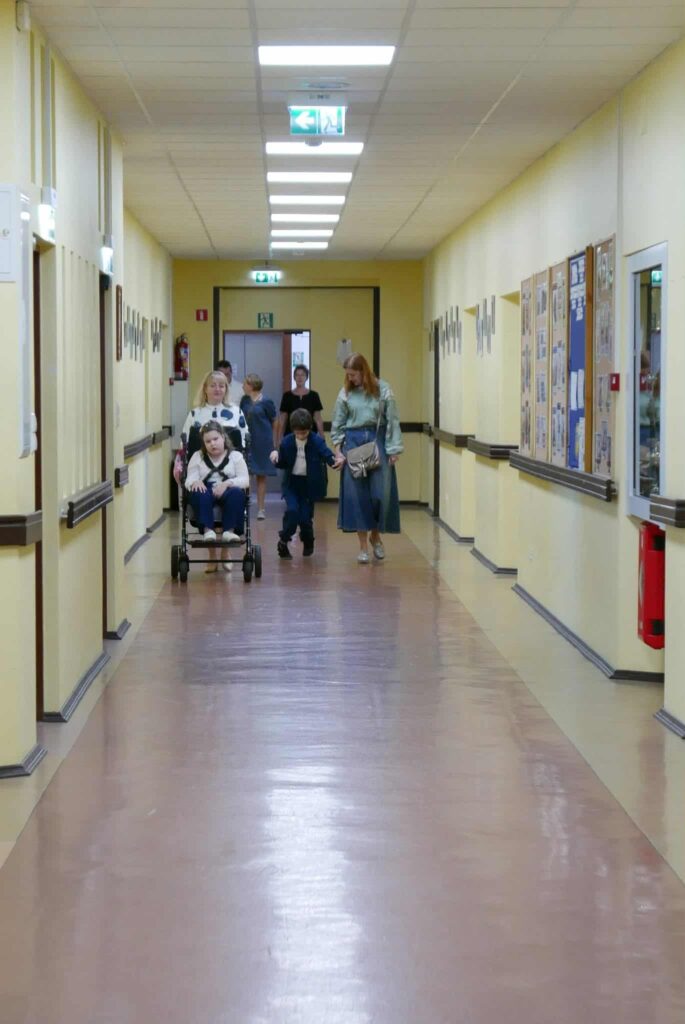 Dzieci i dorośli, przemieszczają się korytarzem szkoły. Na czele grupa z wózkiem inwalidzkim, w którym siedzi dziecko. Obok idą kobieta i chłopiec trzymający się za ręce.