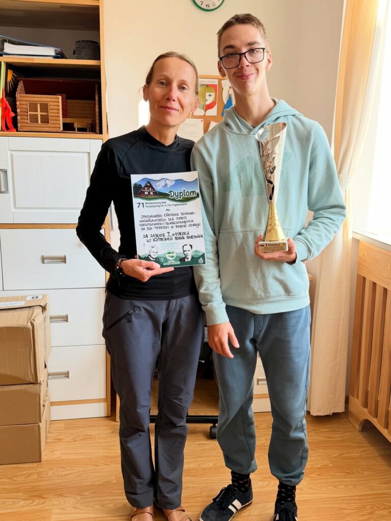 Chłopiec w okularach trzyma w ręce puchar, obok stoi nauczycielka trzymając w ręce dyplom.
