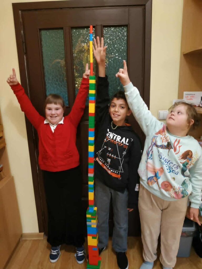 Dwie dziewczynki i chłopiec stoją przed wysoką wieżą z klocków. Uśmiechają się i mają uniesione rece.