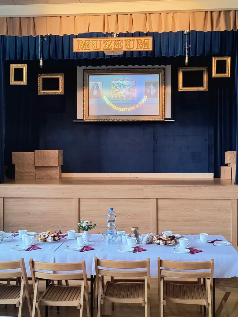 Scena na górze widać wiszące dwa mikrofony oraz napis Muzeum. Z tyłu na ekranie w ramie zdjęcia dwóch zakonnic i napis "Wspomnień czas". Przed sceną zastawiony stół.