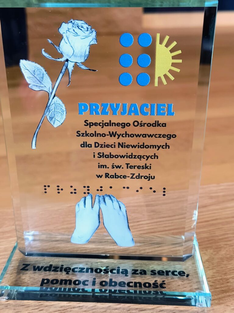 Szklana statuetka z logo SOSW w Rabce - Zdroju z prawej strony i różą z lewej oraz napis "Przyjaciel SOSW dla Dzieci Niewidomych i Słabowidzących im.św Tereski w Rabce-Zdroju. pod spodem napis w brajlu i dłonie. Na podstawie napis " Z wdzięcznością za serce, pomoc i obecność"