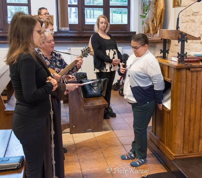 Młoda dziewczyna w okularach stoi przy mównicy w kaplicy w trakcie Mszy św. i trzyma w reku mikrofon. naprzeciwko niej stoją 3 kobiety, z których jedna gra na gitarze.