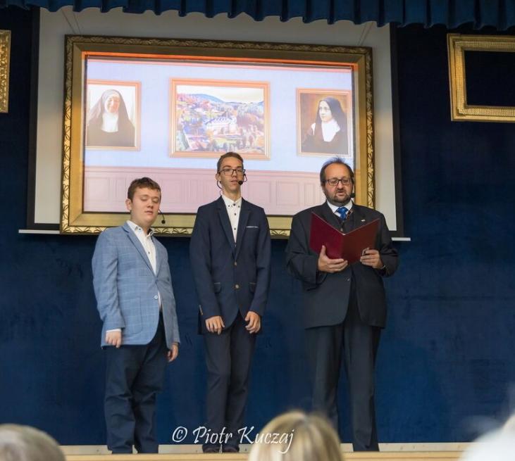 Trzech mężczyzn stoi na scenie, mężczyzna po prawej czyta. w tle duży obraz ze zdjęciami dwóch zakonnic i widokiem Szkoły im. św. Tereski w Rabce. 