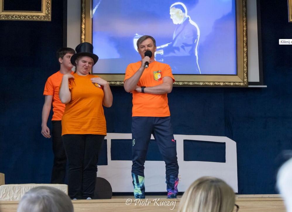 Trzy osoby w pomarańczowych koszulkach stoją na scenie; jedna z nich trzyma mikrofon i mówi, inna ma na głowie cylinder. W tle widać ekran z projekcją postaci.