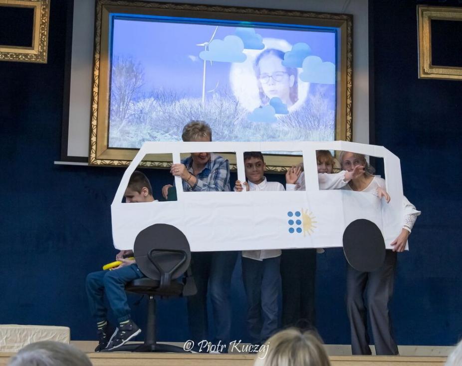 Grupa osób stoi na scenie, trzymając dużą makietę samochodu z papieru. W środku „pojazdu” widać uczestników machających do publiczności.
