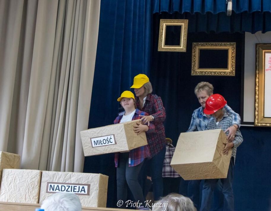 Osoby w kaskach i koszulach w kratę stoją na scenie, trzymając duże kartonowe pudełka z napisami „MIŁOŚĆ” i „NADZIEJA”.