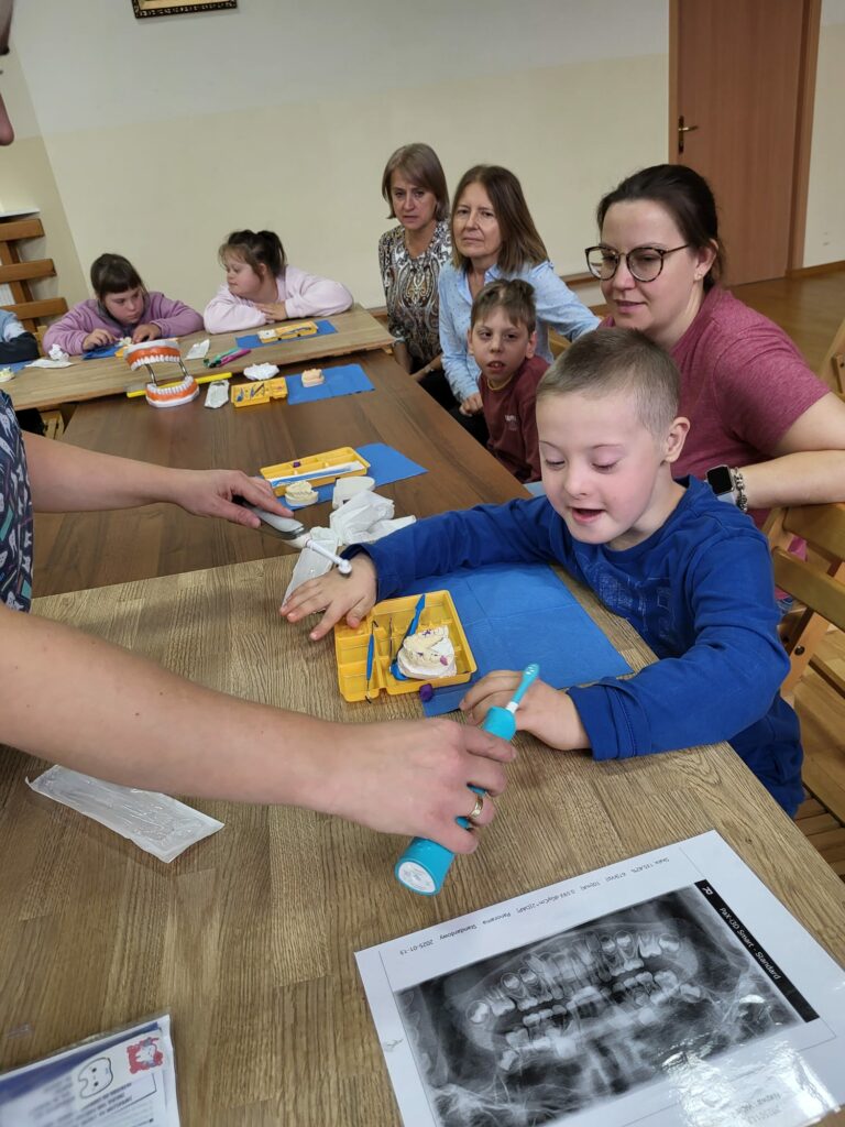 Grupa osób podczas warsztatów stomatologicznych siedzi przy stołach. Chłopiec testuje na dłoniach działanie szczoteczek elektrycznych.