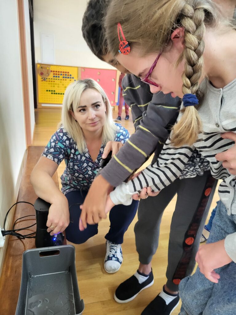 Dziewczynka w okularach i chłopiec podają ręce prowadzącej warsztaty, nachylając się nad miską z wodą.