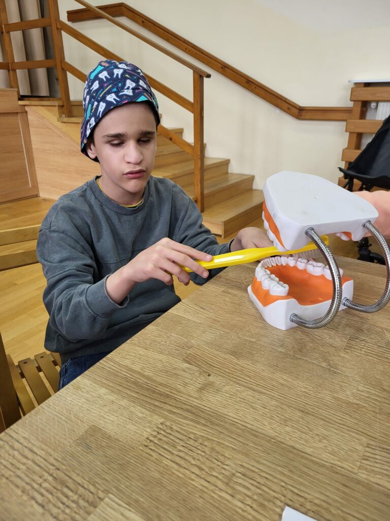 Chłopiec w kolorowej czapce uczy się prawidłowo szczotkować zęby na dużym modelu szczęki