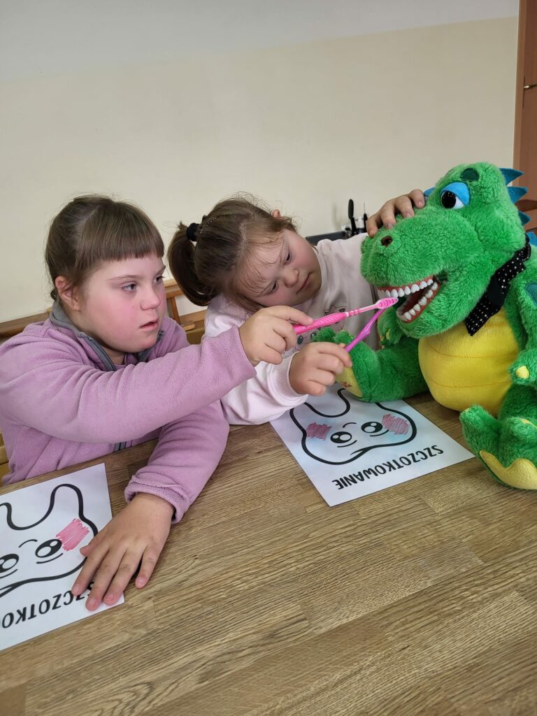 Dwie dziewczynki ćwiczą szczotkowanie zębów na  maskotce zielono - żółtego smoka.