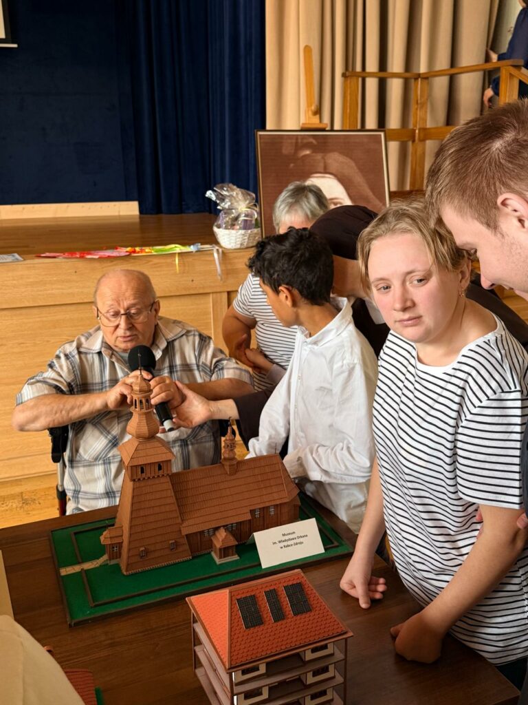 Grupa osób ogląda makietę drewnianego kościoła, którą prezentuje starszy mężczyzna na wózku. W tle scena i portret kobiety.