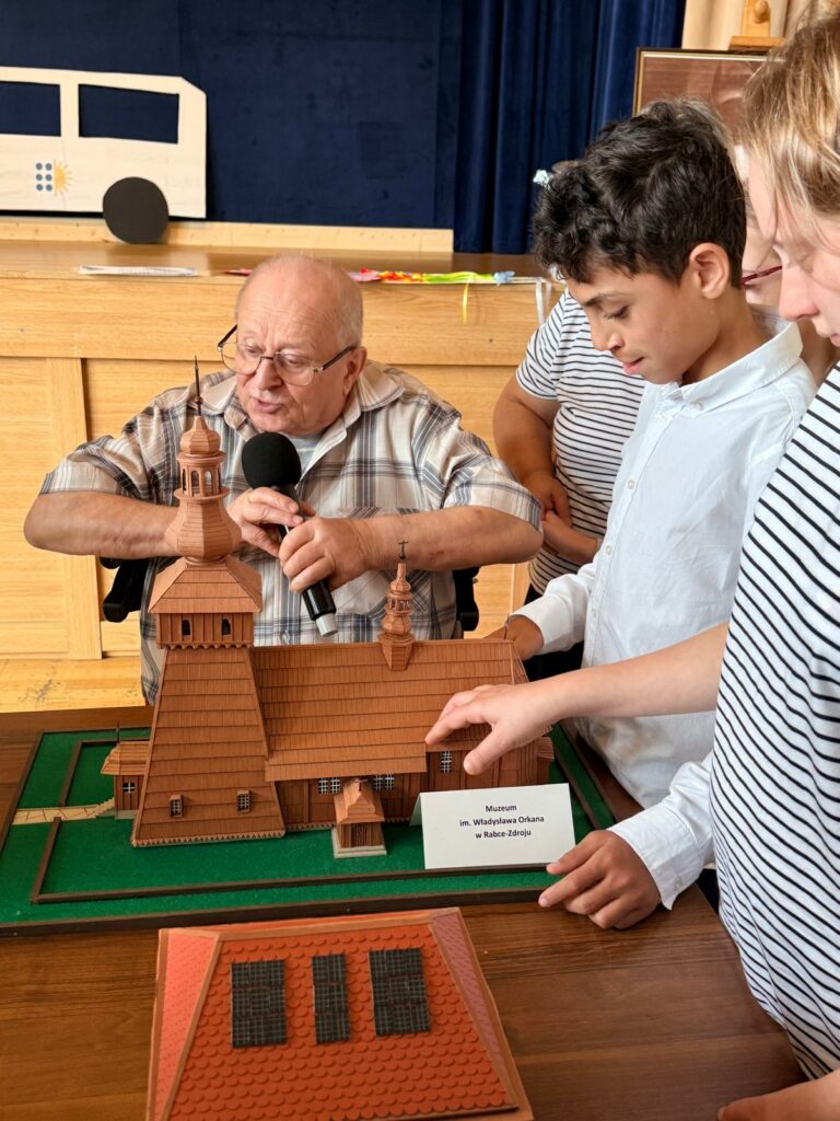 Dziewczyna i chłopiec oglądają makietę drewnianego kościoła, którą prezentuje starszy mężczyzna na wózku.