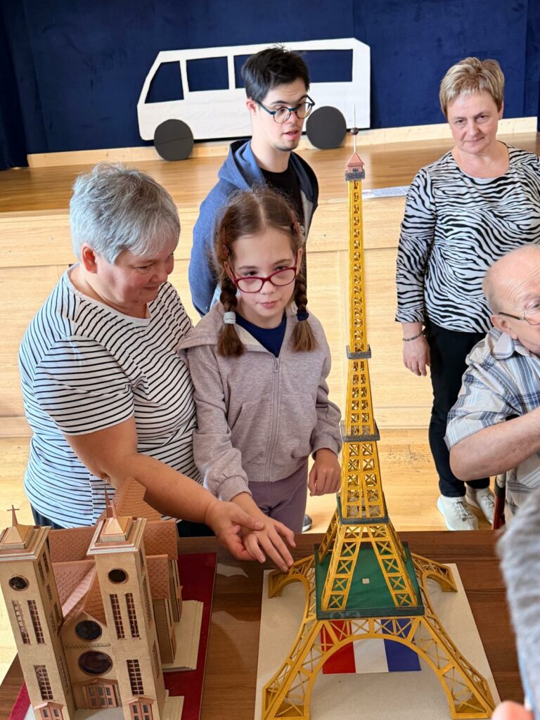Dziewczynka dotyka makiety wieży Eiffla, prowadzona przez kobietę. Obok znajdują się inne osoby oraz model katedry.