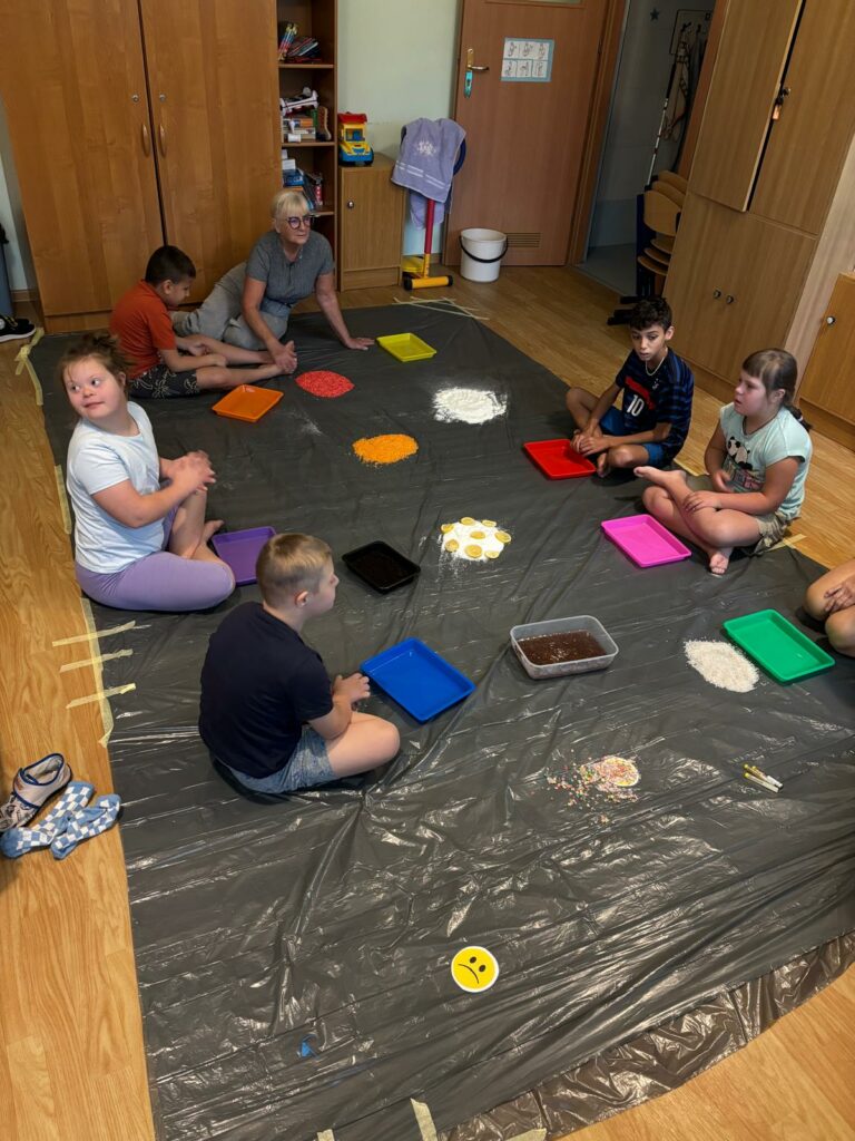 Grupa dzieci i nauczycielka siedzą w kręgu na podłodze, na rozłożonej folii z rozsypanymi kolorowymi materiałami i tackami przed sobą.