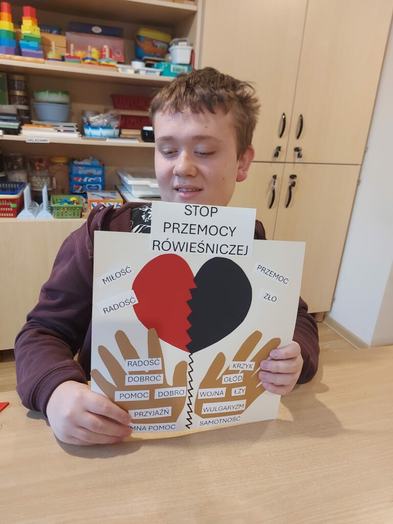 Chłopiec trzyma plakat z hasłem „STOP PRZEMOCY RÓWIEŚNICZEJ”. Na plakacie widnieje pęknięte serce, dłonie oraz słowa symbolizujące dobro (np. „miłość”, „przyjaźń”) i zło (np. „przemoc”, „krzyk”).