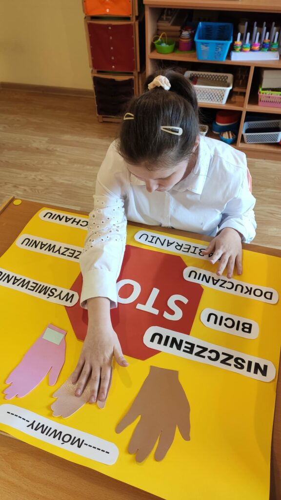 Dziewczyna w białej bluzce nakleja na duży plakat wyciętą dłoń. na środku znajduje się duży znak stop otoczony napisami.