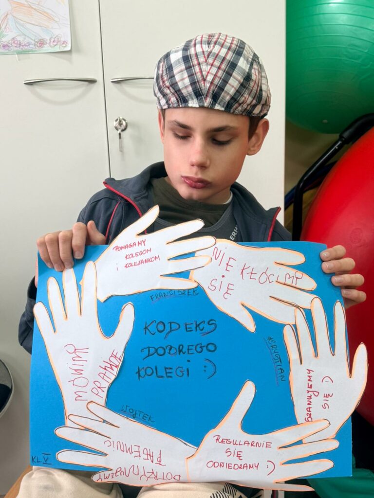 Chłopiec trzyma w rękach pracę plastyczną. Na niebieskim tle naklejone białe ręce z napisami, na środku napis "Kodeks dobrego kolegi"