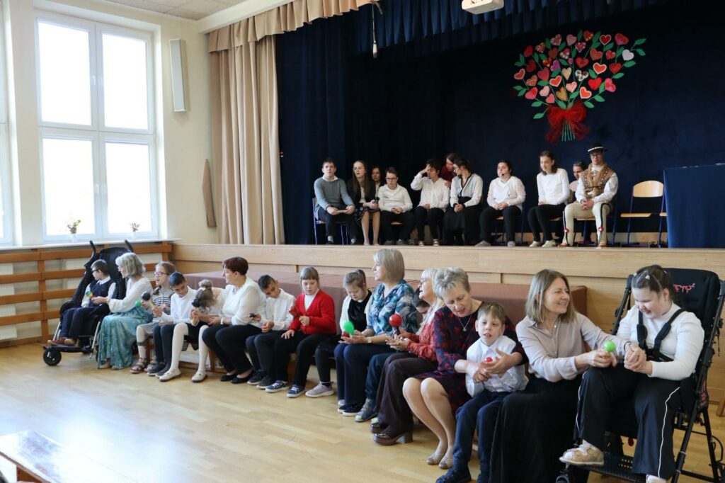 Grupa osób siedzi na scenie, a druga pod sceną gra na różnych instrumentach.