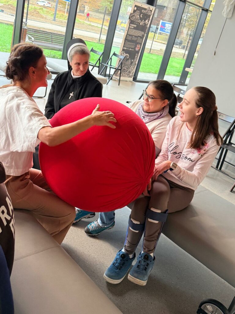 Instruktorka pokazuje dużą czerwoną piłkę owiniętą materiałem dwóm dziewczynkom i siostrze zakonnej, wszyscy uśmiechnięci, siedzą na ławce.